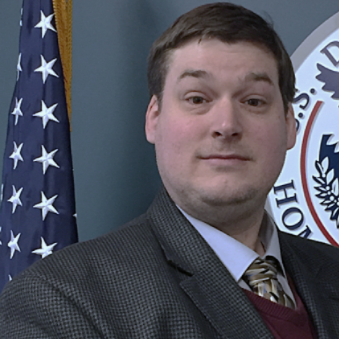 Profile Picture for Michael Kane standing in front of a flag on a pole, and a USCIS emblem.